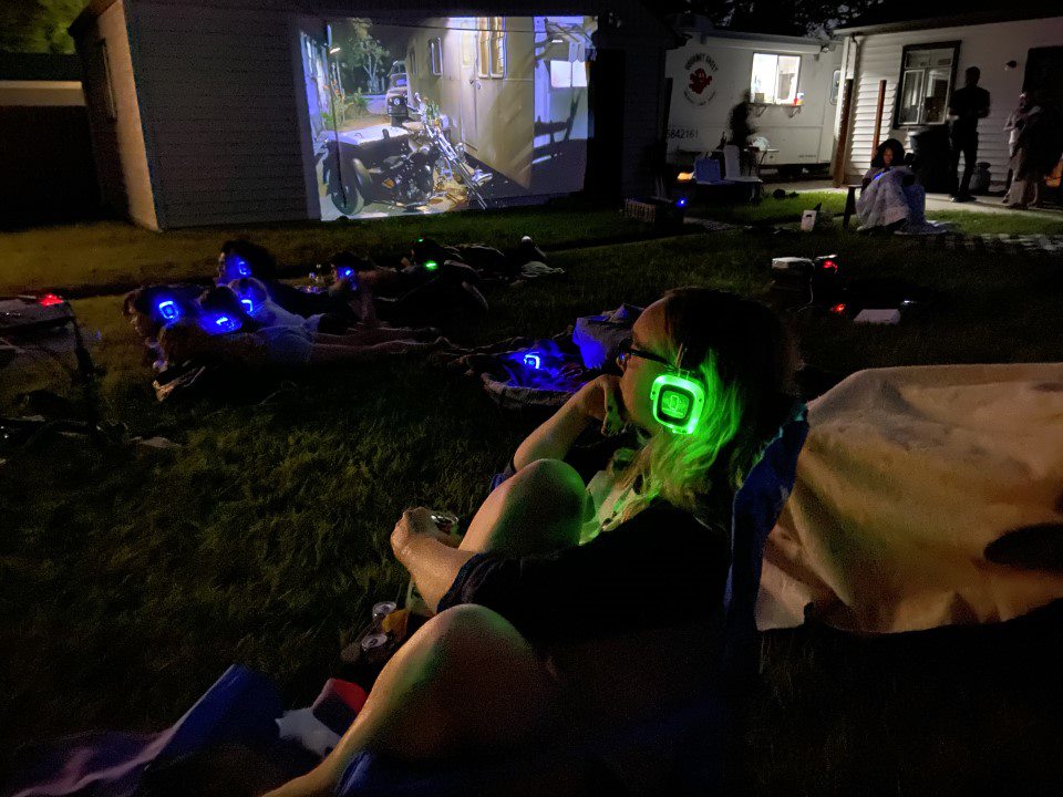 crowd wearing glowing silent disco headphones facing an outdoor movie screen at dusk.